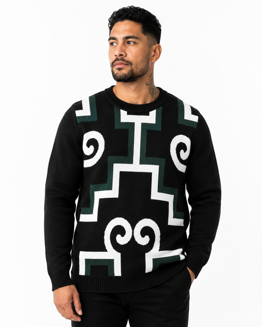 A man wearing a black crewneck sweater with a bold white and dark green geometric tribal pattern, set against a plain white studio background.