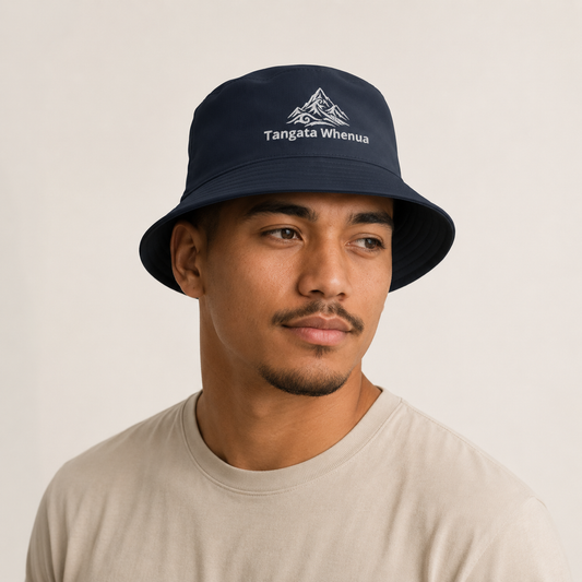 A man wearing a navy blue bucket hat with a white embroidered mountain graphic and the text 'Tangata Whenua' on the front, paired with a light beige t-shirt against a plain off-white background.
