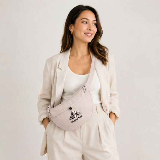 A woman wearing a light beige blazer, white tank top, and matching trousers models a light pink canvas crossbody bag with a black mountain logo and the text 'Tangata Tiriti'.