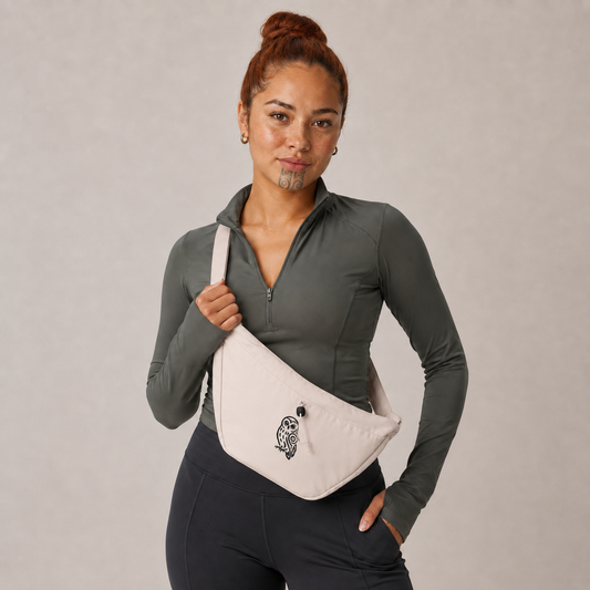 A woman wearing a dark gray long-sleeve athletic top and black leggings models a light beige crossbody fanny pack with a black embroidered logo, set against a neutral gray studio background.