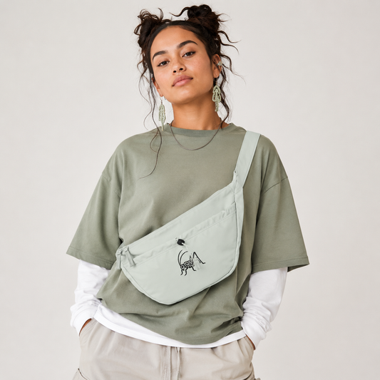 A woman wearing an oversized sage green t-shirt, a white long-sleeve shirt, and beige pants, carrying a light sage green crossbody bag with a black embroidered wētā design on the front pocket.