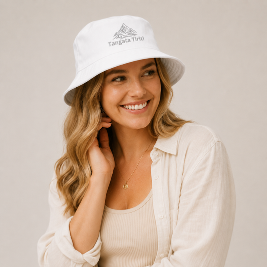 A smiling woman wearing a white bucket hat with a gray mountain logo and the text 'Tangata Tiriti' on the front, paired with a cream-colored button-up shirt against a neutral beige background.