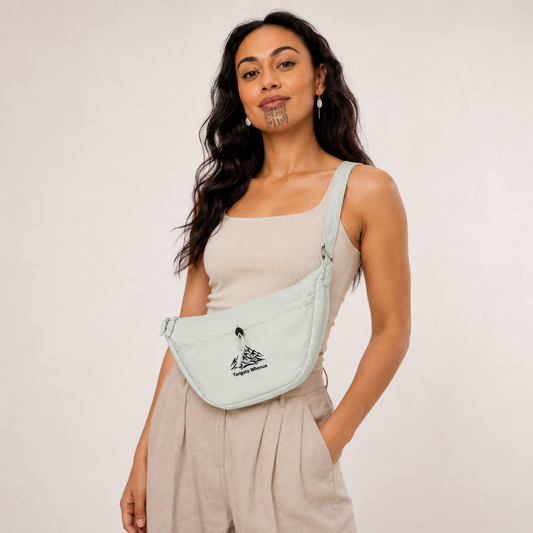 A woman wearing a light sage green sling bag with a black mountain logo and the text 'Tangata Whenua' across the front, styled with a beige ribbed tank top and beige trousers against a plain off-white background.